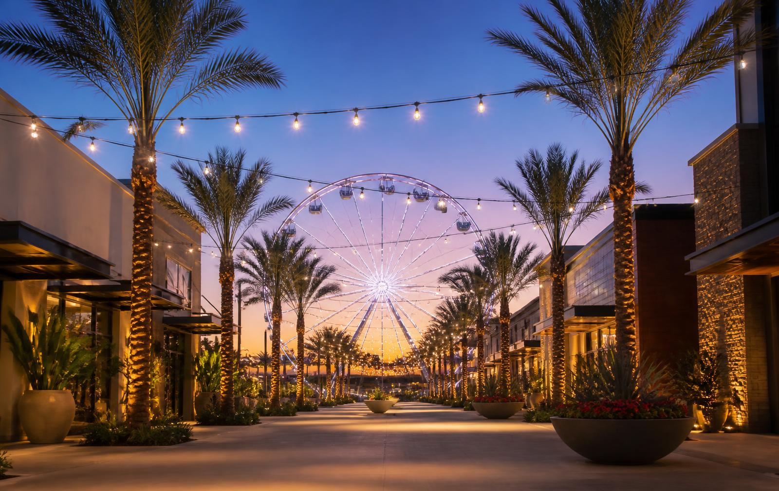 Outdoor Southern California shopping promenade at dusk with string lights, palm trees and an illuminated observation wheel